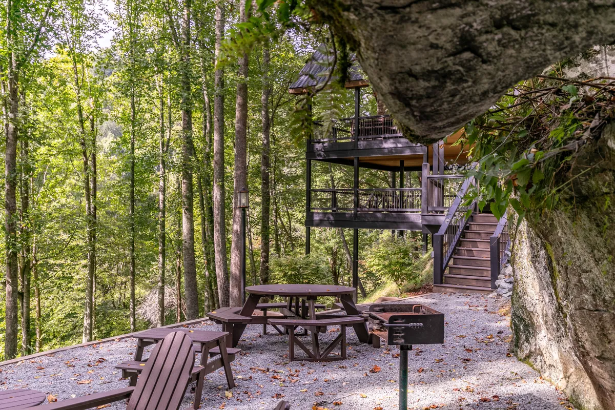 A multi-level wooden deck featuring a picnic table and Adirondack chairs, surrounded by tall trees. The setting is cozy and encourages outdoor activities like dining, relaxing by the fire pit, and enjoying the natural environment.