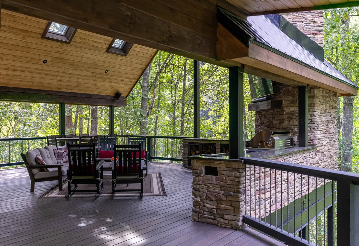 An inviting exterior seating area with a stone fireplace, grill, and comfortable chairs, surrounded by lush greenery. The space encourages socializing, outdoor meals, and relaxing in a tranquil natural environment.
