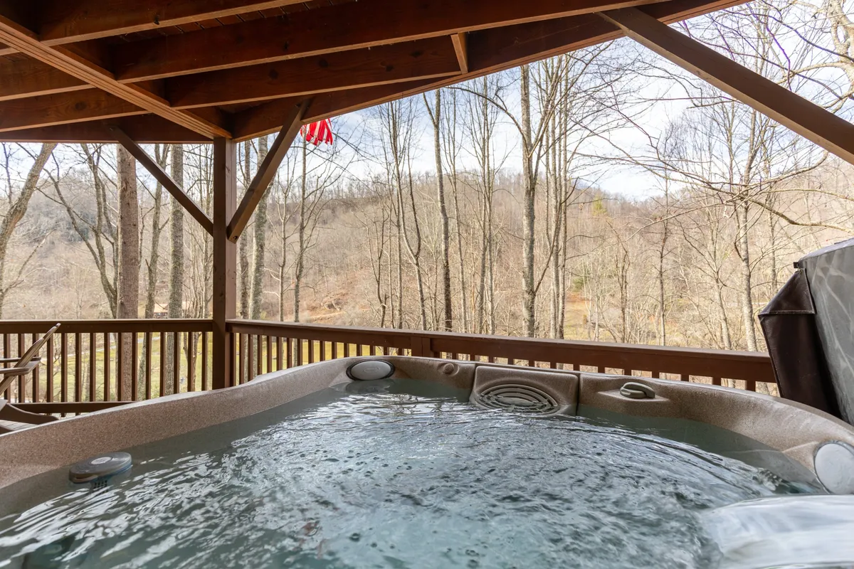 An outdoor hot tub sits on a wooden deck overlooking lush trees, with the American flag fluttering nearby. The scene suggests relaxation and soaking while surrounded by nature—a perfect way to cap off any mountain adventure, regardless of the season.
