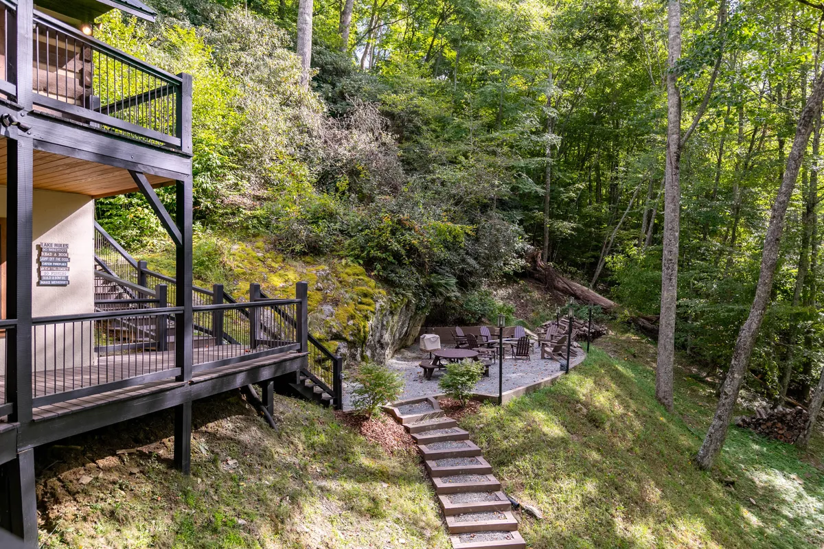 A spacious deck with railing overlooks a stone-paved patio dotted with wooden chairs, all set amidst lush greenery. The setting is ideal for outdoor dining, morning coffee, or gathering with friends and family before or after local outings.