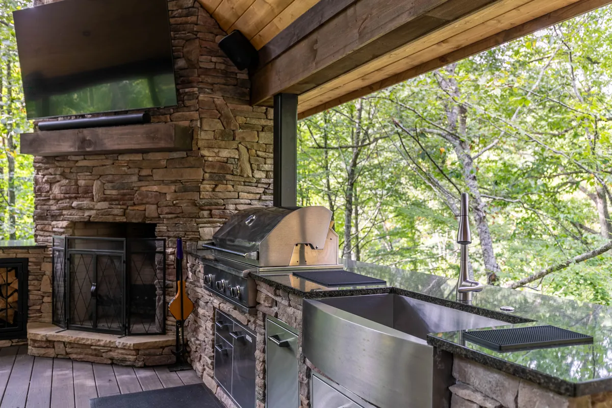This image features a spacious outdoor kitchen with a stone fireplace, a large TV, and natural scenery in the background. The setup is perfect for entertaining, grilling, and enjoying meals together while watching a game or relaxing by the fire.