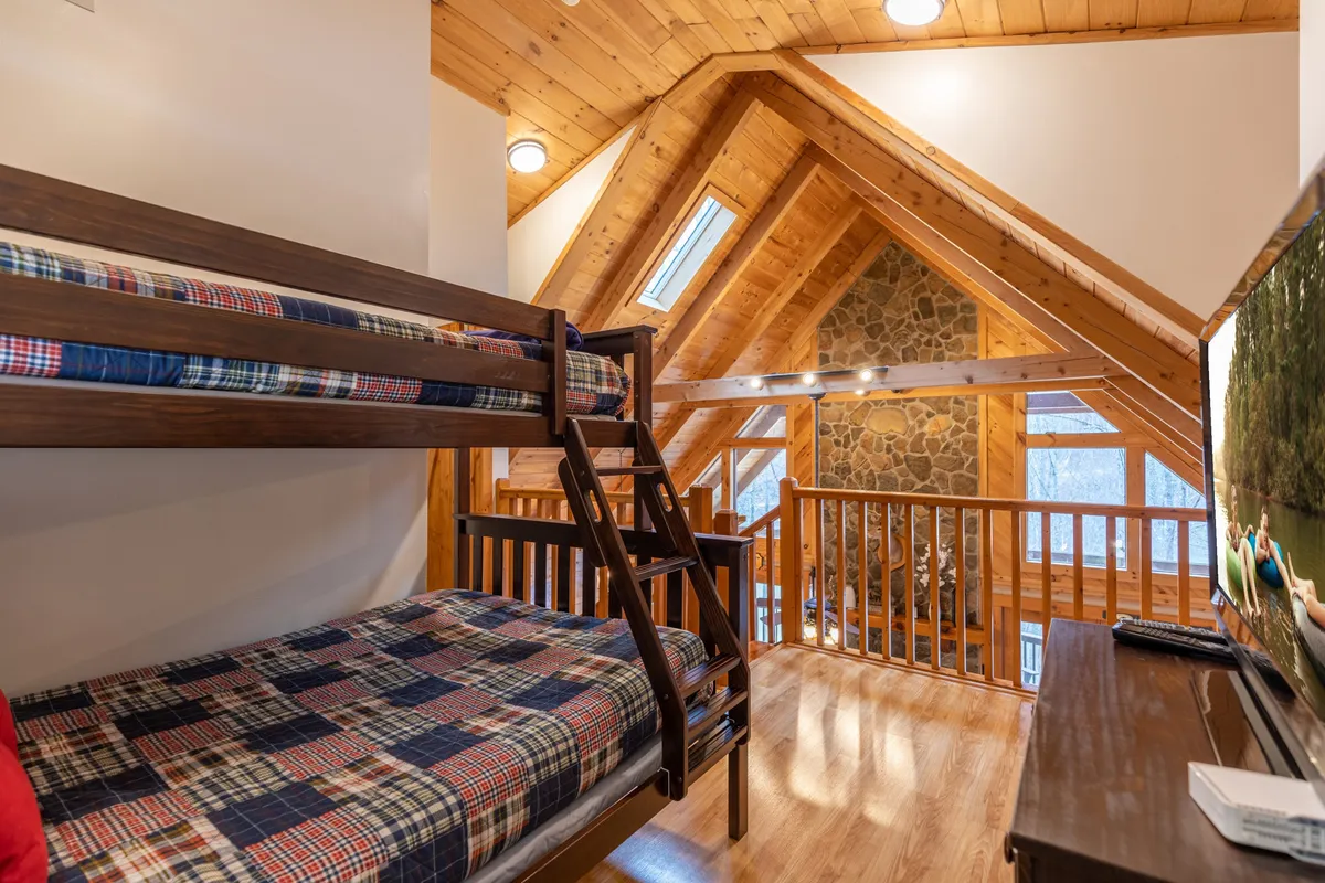 An airy upstairs loft area featuring charming bunk beds, a rustic stone wall, expansive windows, and wooden railings. The bright, open space is designed for younger guests to play, read, or unwind in a safe, cheerful environment.