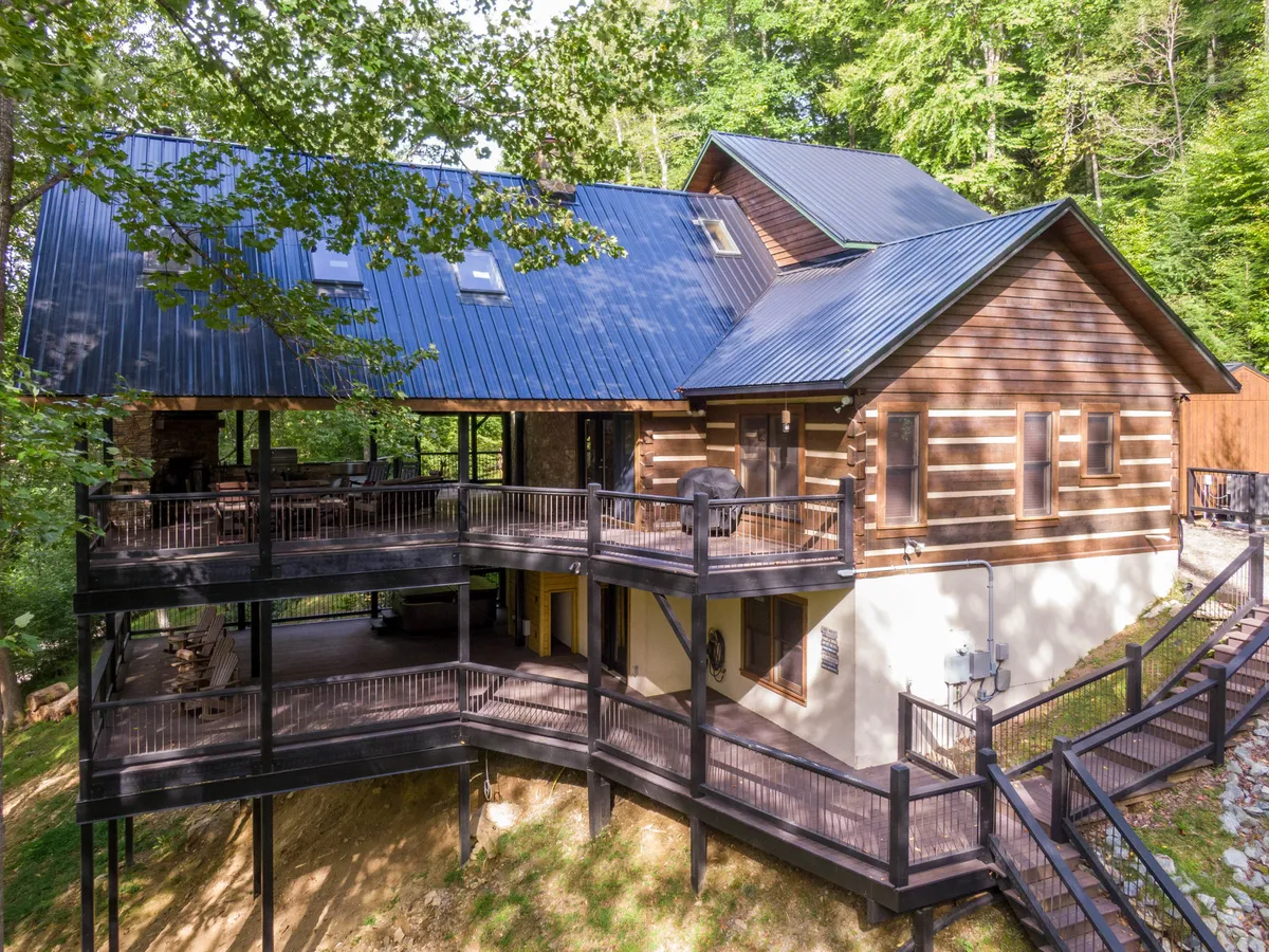 This image features a log cabin structure with an expansive deck surrounded by a dense, green forest. The deck offers ample space for gatherings and relaxing outdoors, while the woodland setting emphasizes the property's close connection to nature and adventure.