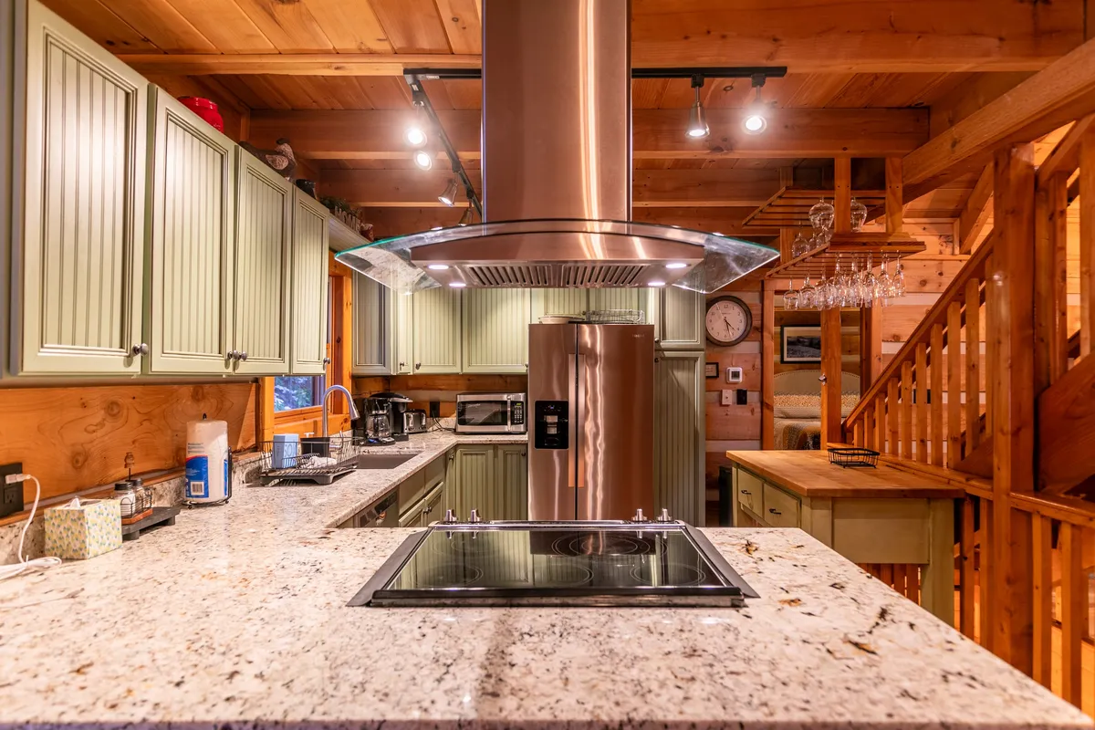 A stylish, fully equipped kitchen with granite countertops, stainless steel range hood, and vibrant green cabinetry. The open layout flows toward a wooden staircase, suggesting easy movement between living, dining, and kitchen zones. The kitchen is staged for cooking and socializing, with plenty of space for guests.