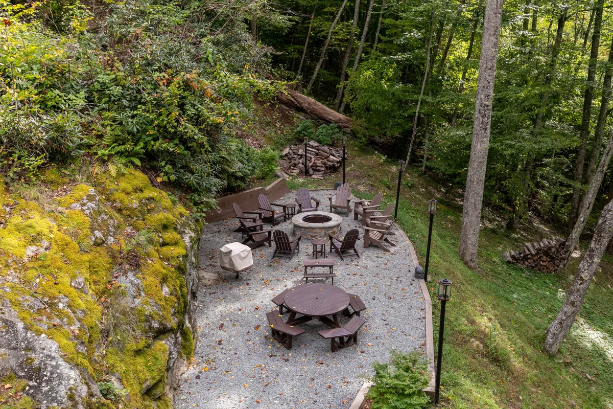 This exterior image showcases a large fire pit surrounded by wooden chairs and a picnic table, set beneath tall trees. The natural setting and inviting seating make it ideal for group gatherings, outdoor dining, and enjoying s’mores or stories by the fire.