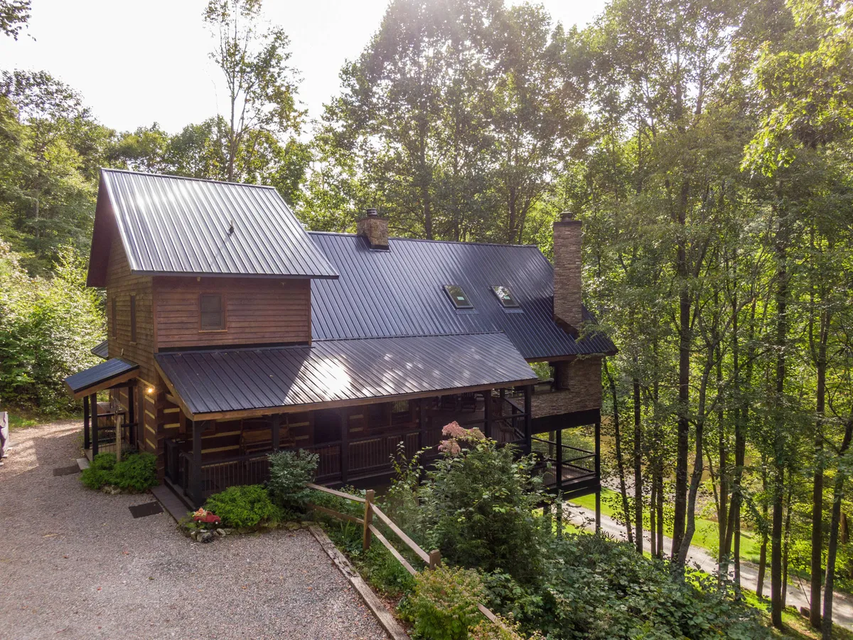 A serene log cabin with a metal roof nestles among mature trees, featuring a welcoming deck area that invites guests to relax. The natural wood construction and surrounding greenery capture the rustic charm and tranquility of the property, setting the scene for mountain adventures.