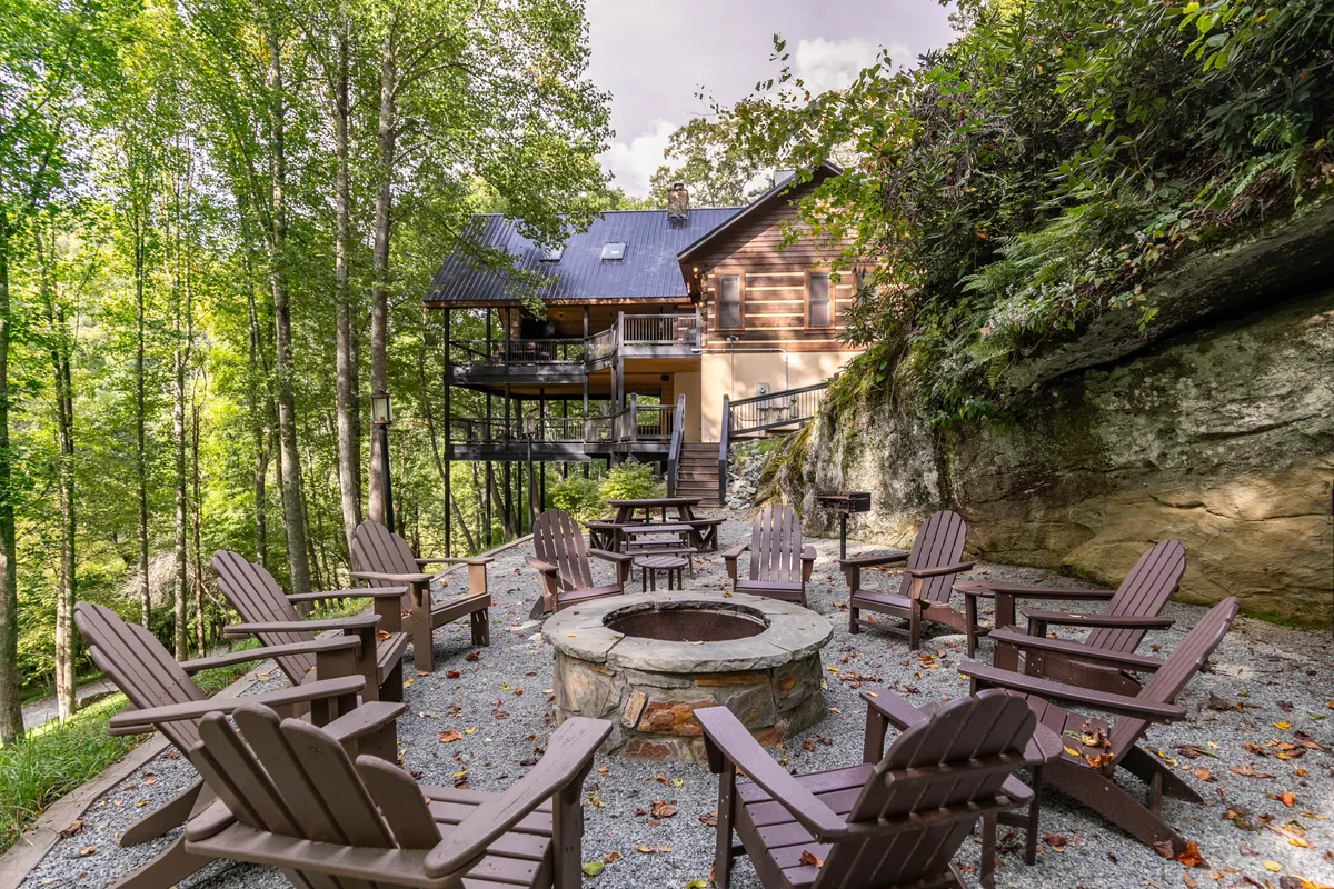 An inviting fire pit circled by classic Adirondack chairs sits just beyond the deck, bordered by tall trees and nature. The scene captures the essence of gathering outdoors—ideal for storytelling, roasting marshmallows, or simply enjoying a starlit evening.
