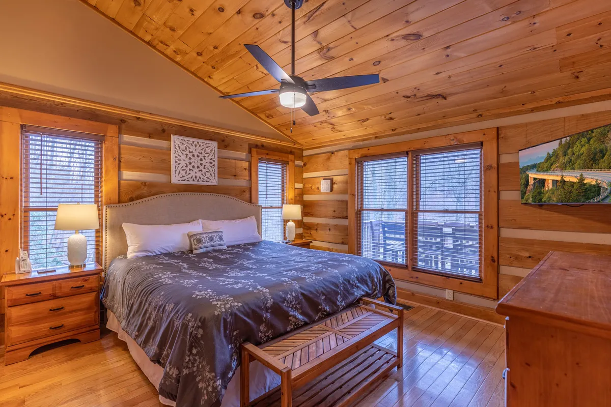 A spacious king-sized bed with inviting wooden furniture sits beside large windows overlooking the natural landscape. The room is accented by a ceiling fan and decorative wall art, creating an atmosphere of comfort and seclusion.