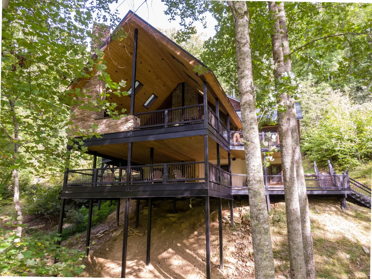 The image showcases the cabin's striking exterior with a spacious wooden deck overlooking the tranquil lake, a prominent stone chimney, sloped roofline, and is surrounded by lush trees, creating a serene and inviting setting for outdoor gatherings.