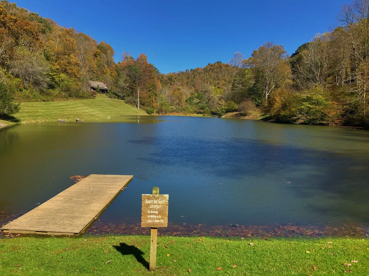 A tranquil lakeside view featuring a wooden dock extending over calm water, surrounded by vibrant autumn foliage. The peacefulness of the lake, with its easy access, is front and center, inviting fishing, nature walks, and lakeside leisure.