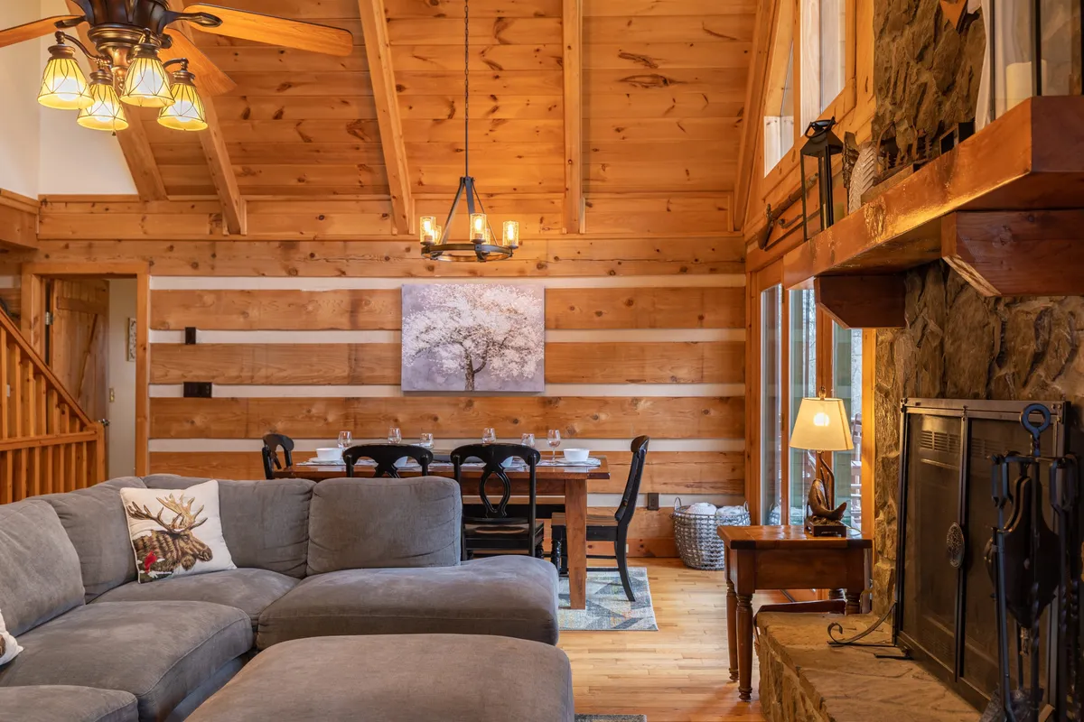 A spacious and inviting interior featuring a large sectional sofa positioned near a stone fireplace, with a dining table and chairs in the background. The artwork and soft lighting create a warm, welcoming atmosphere ideal for gatherings, relaxation, and sharing meals.