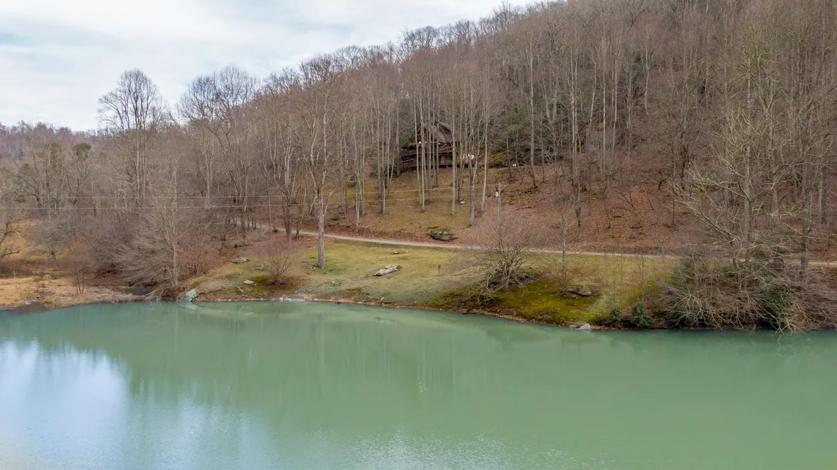 This image captures a tranquil view of the lake with the wooded hillside and the cabin perched nearby. The scene emphasizes the close proximity of the water to the home, highlighting opportunities for fishing, canoeing, and relaxing by the lake's edge.